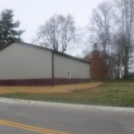 A large metal-sided pole barn with a maroon base sits next to a two-story brick house, both surrounded by leafless trees and grass, beside a paved road under a cloudy sky. Kentucky Builders And Excavating