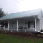 A white house building with a metal roof and a covered front porch featuring chairs, tables, and hanging flower baskets. The house sits on a grassy lawn with trees in the background on a foggy day. Kentucky Builders And Excavating