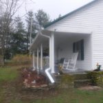 A white building with a covered porch featuring a white rocking chair. The yard has grass, trees, and a small garden bed lined with stone near the porch. The overcast sky adds charm to this peaceful setting. Kentucky Builders And Excavating