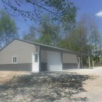 A large metal pole barn with two white doors and a sloped roof, set on a gravel driveway and surrounded by trees on a sunny day. Kentucky Builders And Excavating
