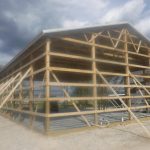 A wooden frame structure for a pole barn is under construction on a gravel lot, with support beams in place beneath a cloudy sky. The open building shows no walls installed yet, and signs of recent excavating are visible around the site. Kentucky Builders And Excavating