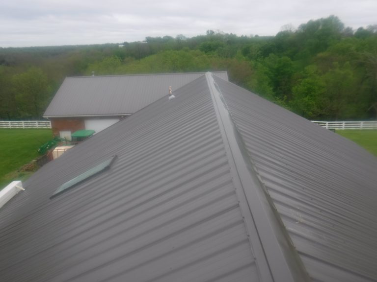 View from atop a dark gray metal roof with a skylight, facing a similar pole barn building and surrounded by green trees and grass under a cloudy sky. White fences border the property. Kentucky Builders And Excavating