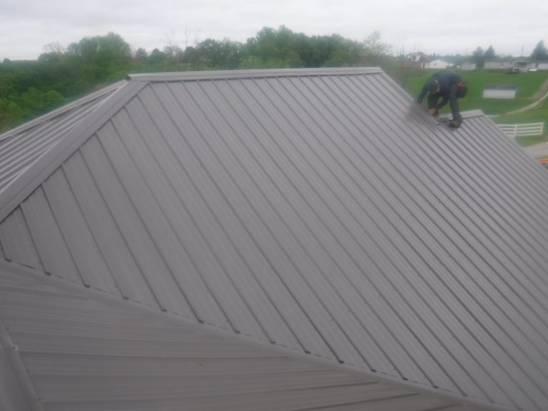 A person crouches near the top edge of a large, gray metal pole barn roof on a cloudy day, surrounded by green fields and distant trees. Kentucky Builders And Excavating