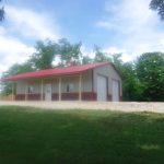 A small metal pole barn with a red roof and cream-and-red siding sits on a gravel lot surrounded by green grass and trees, under a partly cloudy sky. Kentucky Builders And Excavating