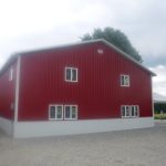 A large red pole barn with white trim and several windows sits on a gravel lot, with trees and a grassy field in the background under a cloudy sky. Kentucky Builders And Excavating