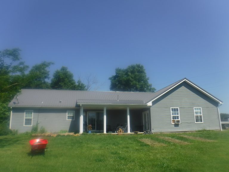 A single-story gray building with a metal roof, front porch supported by columns, and a green lawn. A red wheelbarrow sits on the grass, with tall trees in the background under a clear blue sky. Kentucky Builders And Excavating