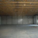 An empty, dimly lit pole barn with metal walls and a wooden beam ceiling. The concrete floor is bare, except for a single red object near the right wall by a small window. Kentucky Builders And Excavating