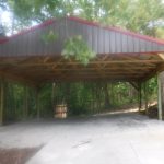 A large open-sided metal and wood pole barn stands on a gravel and concrete pad in a wooded area, surrounded by trees and greenery. Kentucky Builders And Excavating