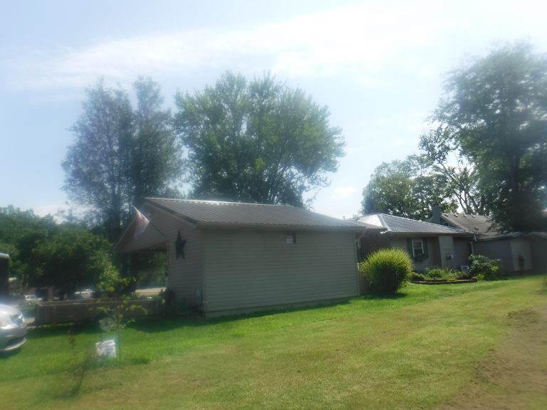 A small house with a metal roof sits on a grassy yard, flanked by trees and plants on a sunny day. Nearby, another building and more greenery are visible in the background. The image appears slightly blurry or hazy. Kentucky Builders And Excavating