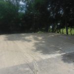 A leveled area of fresh gravel or sand, possibly prepped for a new building or pole barn, adjoins a cracked concrete surface bordered by trees casting shadows, with a grassy field visible through the trees in the background. Kentucky Builders And Excavating