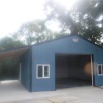 A blue metal pole barn garage with white-trimmed windows and a large open door. A covered carport extends from one side. The building sits on a concrete driveway surrounded by trees, with sunlight filtering through the branches above. Kentucky Builders And Excavating