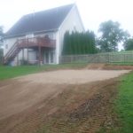 A backyard with a large, leveled patch of dirt and tire tracks from excavating for a pole barn, next to a white house with a red deck and a fenced yard; trees and grass surround the area. Kentucky Builders And Excavating