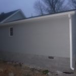 A newly built extension with light gray brick walls, a small window, and white gutters is attached to a house at dusk. The ground around the buildings foundation shows signs of recent excavating. Leafless trees are visible in the background. Kentucky Builders And Excavating