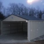 A beige metal pole barn carport with an open front stands on gravel at dusk, surrounded by bare trees and a grassy area, with raindrops visible on the camera lens. Kentucky Builders And Excavating