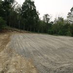 A freshly leveled gravel area, ideal for building a pole barn, sits surrounded by trees. Visible tire tracks cross the surface, and a sloped edge of exposed soil on the left hints at recent excavating work. Kentucky Builders And Excavating