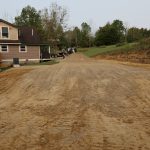A newly leveled dirt driveway extends from a house on the left toward vehicles, a pole barn, and trees in the distance, with grassy banks and earth on either side under a clear sky. Kentucky Builders And Excavating