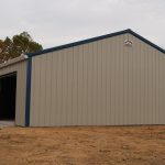 A large beige metal pole barn with blue trim stands on bare ground, featuring an open garage door and a few windows, surrounded by trees under a cloudy sky. Signs of recent excavating are visible around the building. Kentucky Builders And Excavating