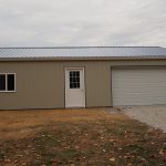 A beige metal pole barn building with a blue-gray roof, featuring a white door, a window on the left, and a large white garage door on the right, sits on bare ground with some grass and leaves. Ideal for storage or excavating projects. Kentucky Builders And Excavating