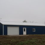 A large blue metal pole barn with a white garage door, a white entry door, and a single window on the front sits on a grassy plot with trees in the background under an overcast sky. Kentucky Builders And Excavating