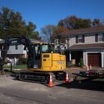 A yellow excavator is parked on a residential street in front of a house with brown garage doors, surrounded by orange traffic cones and a trailer—ready for excavating work. Trees, other houses, and a clear blue sky complete the scene. Kentucky Builders And Excavating