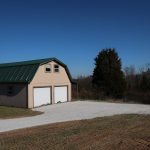 A beige pole barn with a green roof and two white garage doors stands beside a gravel driveway, surrounded by trees and open land under a clear blue sky. Kentucky Builders And Excavating