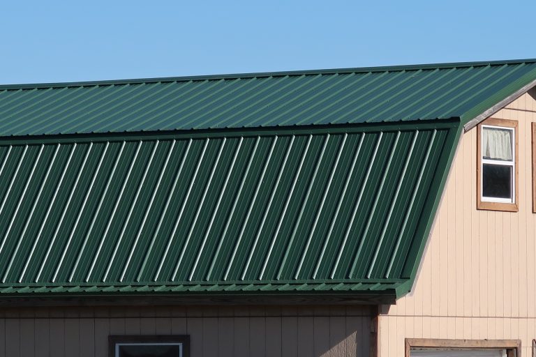 A green metal roof with vertical ridges tops a light tan pole barn with brown trim under a clear blue sky. Two windows are partially visible on the building. Kentucky Builders And Excavating