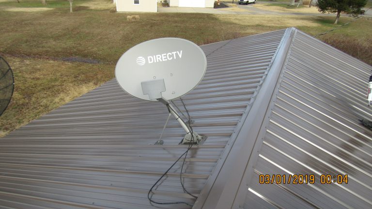 A DirecTV satellite dish is mounted on a gray metal roof with cables running from the dish. The roof, possibly part of a pole barn building, overlooks a grassy yard and nearby houses. The photo is dated 03/01/2019. Kentucky Builders And Excavating