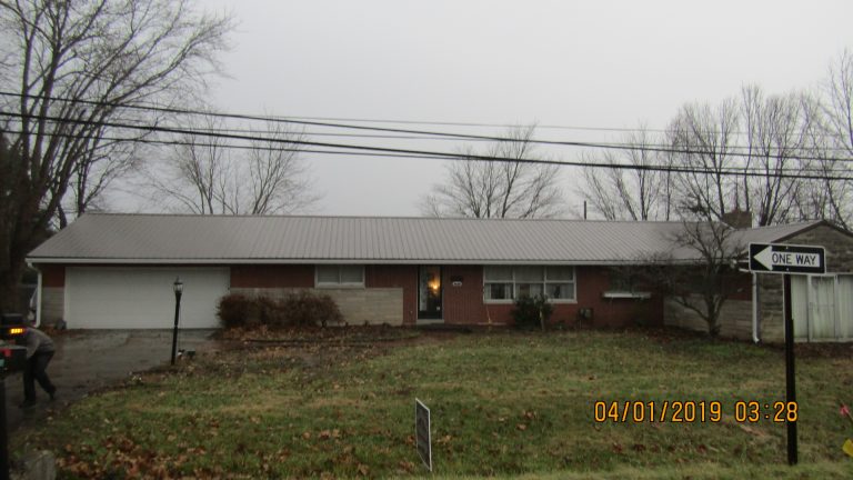 A single-story brick building with a metal roof sits on a grassy lot with leafless trees. A One Way street sign is in front, and a person is walking near the driveway. The photo is dated 04/01/2019. Kentucky Builders And Excavating