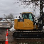 A yellow excavator is parked on a snowy residential street near a house, likely used for building or excavating. Orange safety cones and a white dumpster are in the background, with bare trees and overcast skies hinting at winter. Kentucky Builders And Excavating