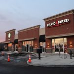 A small shopping plaza at dusk with a Starbucks and a Rapid Fired Pizza. Orange traffic cones block parts of the parking lot as excavating work takes place near the building, and empty outdoor tables are visible near the pizza restaurant. Kentucky Builders And Excavating