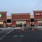 A strip mall building with a Starbucks on the left and Rapid Fired Pizza on the right; the parking lot has orange cones, accessible parking spaces, and signs of recent excavating near the entrances. Kentucky Builders And Excavating