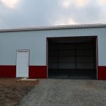 A large metal pole barn with red and white siding features a closed white door on the left and a wide open garage door on the right, revealing an empty interior. The building sits on a gravel and dirt surface, ideal for excavating projects. Kentucky Builders And Excavating