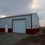 A large white and red metal pole barn with a roll-up garage door and a smaller entry door stands on a gravel driveway, surrounded by open fields and a cloudy sky in the background. Kentucky Builders And Excavating