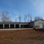 A large gray metal pole barn with multiple garage bays and a single overhead door stands on a dirt lot, with leafless trees in the background under a blue sky. Kentucky Builders And Excavating