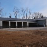 A large gray metal pole barn with five open garage bays and one closed section sits on a gravel lot, surrounded by bare trees under a partly cloudy sky. Kentucky Builders And Excavating