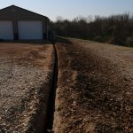 A freshly dug trench runs alongside a gravel driveway near a large garage, perfect for building or pole barn projects, with two white doors and trees set against a clear sky in the background. Kentucky Builders And Excavating