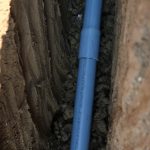 A close-up view of a gray PVC conduit pipe laid in a narrow trench dug into the soil, capturing the surrounding dirt and texture of the trench walls during pole barn excavating. Kentucky Builders And Excavating