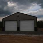 A metal pole barn with two large white garage doors sits on a concrete slab under an overcast sky. The dirt and gravel surrounding the building hint at recent excavating, with trees and greenery in the background. Kentucky Builders And Excavating
