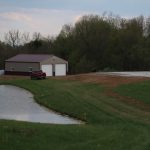A red pickup truck is parked on a grassy area near a pond, next to a tan pole barn with white doors. Trees and a cloudy sky are in the background. A second car is parked near the building. Kentucky Builders And Excavating