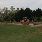 A yellow bulldozer is excavating gravel on a dirt path in a grassy field for a building project, with trees and a cloudy sky in the background. Kentucky Builders And Excavating