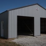 A light gray pole barn storage building with two large open garage doors and a separate side door, situated on a gravel driveway under a blue sky. Perfect for building projects or storing excavating equipment. Kentucky Builders And Excavating