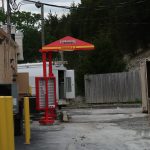 A Firehouse Subs-branded book-sharing box stands near a fenced area in an alley with yellow bollards, a storage container, and building equipment on a cloudy day. Kentucky Builders And Excavating