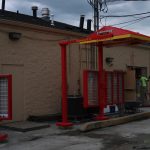 A drive-thru ordering station under construction outside a beige building, with red metal frames, menu boards, and excavating equipment visible. A worker in a green shirt stands near an open door amid construction materials. Kentucky Builders And Excavating