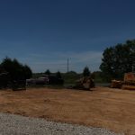 A construction site with a large bulldozer excavating freshly leveled dirt, a dump truck on a gravel area, and trees and grass in the background under a clear blue sky—perfect conditions for building a new pole barn. Kentucky Builders And Excavating