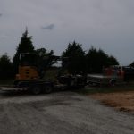 A small excavator, used for building or excavating a pole barn, is loaded onto a trailer hitched to a pickup truck, parked on a gravel road surrounded by trees and open land under a cloudy sky. Kentucky Builders And Excavating