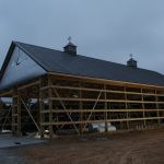 A large wooden pole barn under construction at dusk, illuminated by lights inside. The building features a metal roof with two cupolas and weather vanes, open framework walls, and the ground around is bare dirt and gravel from recent excavating. Kentucky Builders And Excavating