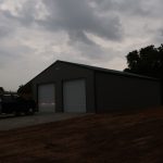 A dark pickup truck is parked in front of a large, gray metal pole barn with two closed garage doors, under a cloudy sky at dusk. Trees and a field are visible in the background. Kentucky Builders And Excavating