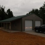 A large metal pole barn with a green roof and two white garage doors stands next to a black pickup truck on a gravel driveway, surrounded by trees and straw-covered ground under an overcast sky. Kentucky Builders And Excavating