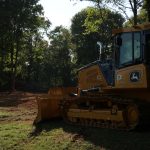 A yellow bulldozer, used for excavating, is parked on a grassy area near the edge of a forest, with sunlight filtering through the trees in the background. Kentucky Builders And Excavating