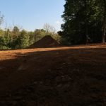 A cleared, dirt-covered area with a mound of soil in the center suggests recent excavating, surrounded by tall green trees under a clear blue sky. Sunlight casts shadows across the ground. Kentucky Builders And Excavating